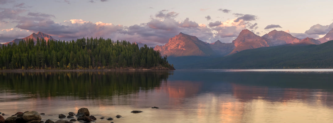 Fine art landscape photograph of Glacier National Park mountains glowing with alpenglow reflected in calm lake water at sunset with evergreen forest shoreline.
