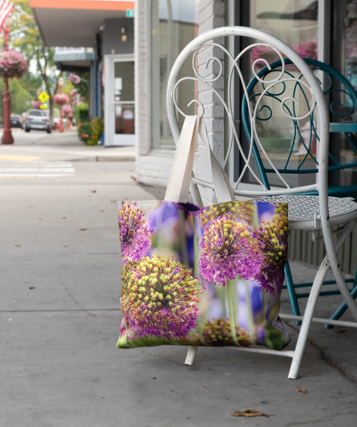 Floral tote bag with purple and yellow allium design hanging on a white metal chair outside a storefront along a sidewalk