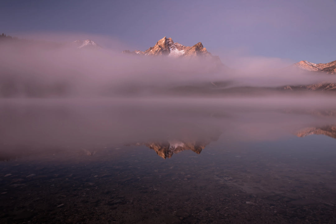 Sawtooth mountain peak emerging through fog with soft reflection in Little Redfish Lake, Idaho at sunrise.