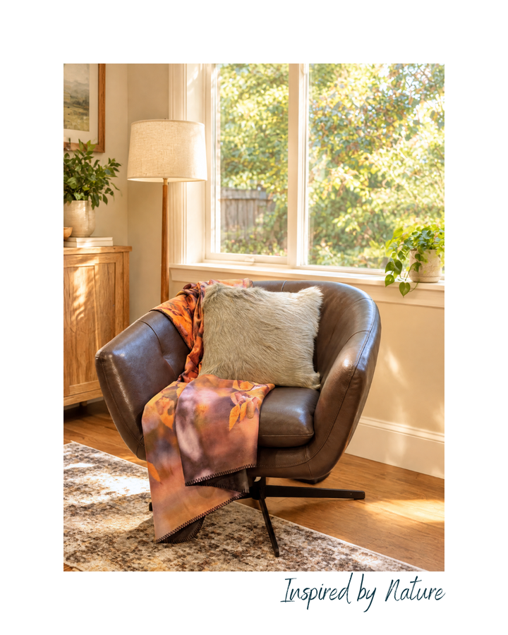 A dark brown leather swivel armchair in a bright, sunlit room with a window, floor lamp, and plants. A patterned orange throw blanket and a textured beige pillow are draped over the chair.