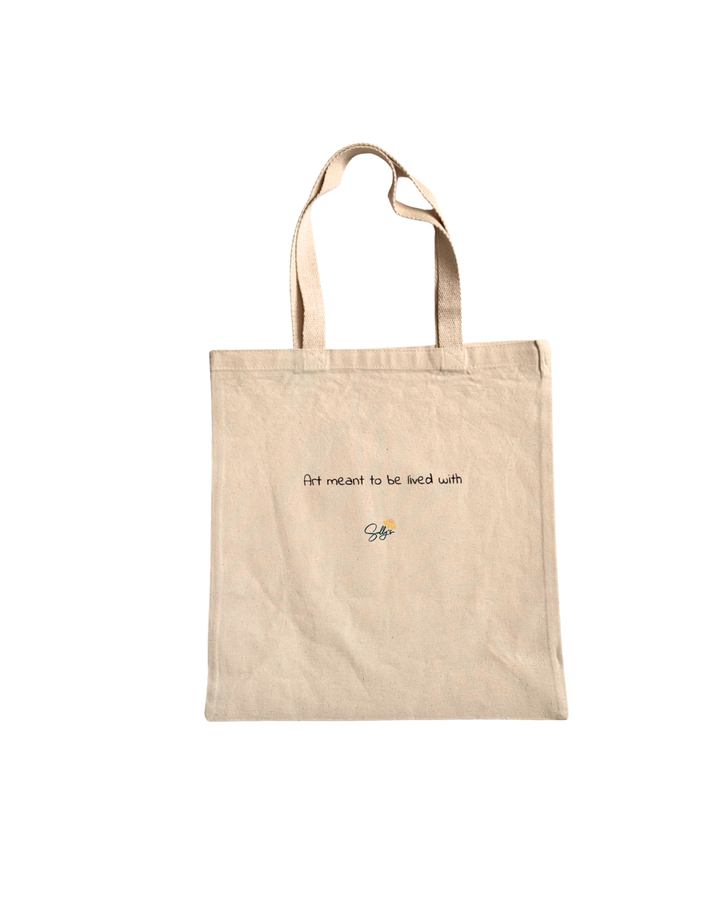 A beige canvas tote bag with two shoulder straps, featuring the printed text 'Art meant to be lived with' and a small colorful logo on the back, set against a plain white background.