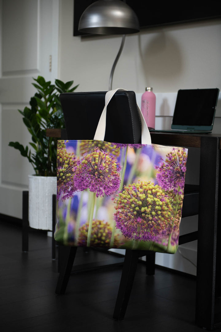 Floral tote bag with purple and yellow allium flower design hanging on a chair at a desk with a laptop, lamp, and potted plant