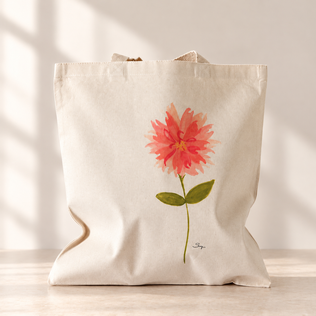 A Little Bit Sassy natural canvas tote featuring a coral watercolor flower with green leaves and Sally Halvorsen’s signature, displayed on a warm neutral tabletop against a cream wall with soft window shadows.