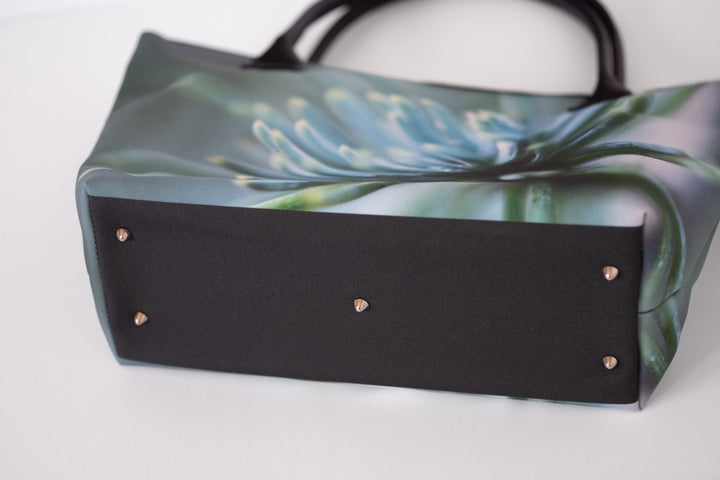 A tote bag with a black fabric base and a printed floral pattern in shades of blue, green, and gray on the top. The bottom edge features a solid black panel with small metallic rivets, and the bag has black shoulder straps, photographed on a white background.