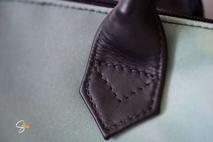 Close-up of a black leather handle attachment with decorative stitching on a light sage green canvas bag.