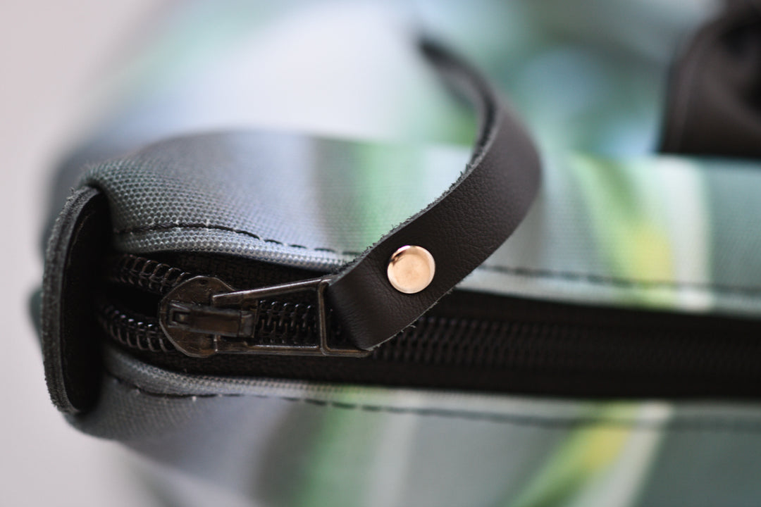 Close-up of a zipper with a black leather strap and a small gold-tone rivet, set against a blurred green and gray patterned background.