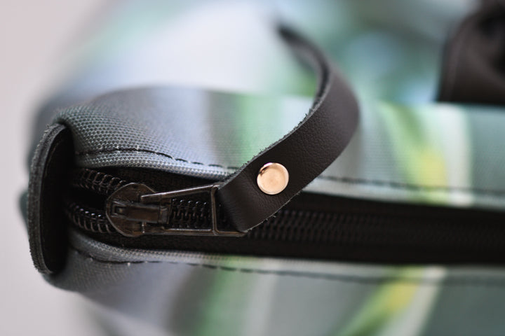 Close-up of a zipper with a black leather strap and a small gold-tone rivet, set against a blurred green and gray patterned background.