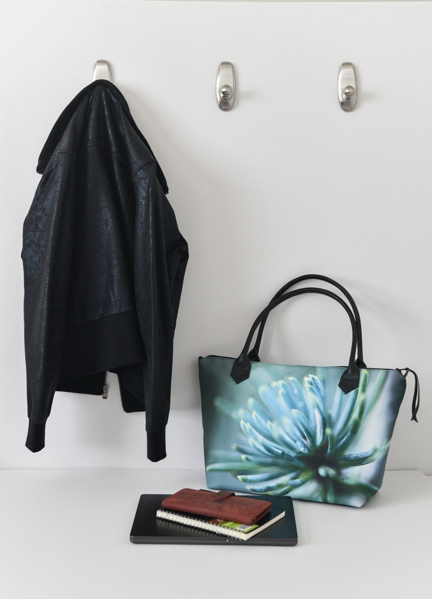 A black faux-leather jacket hangs on a wall hook, next to a tote bag with a blue-green floral print. A small brown wallet and notebook rest on a black computer on the floor.