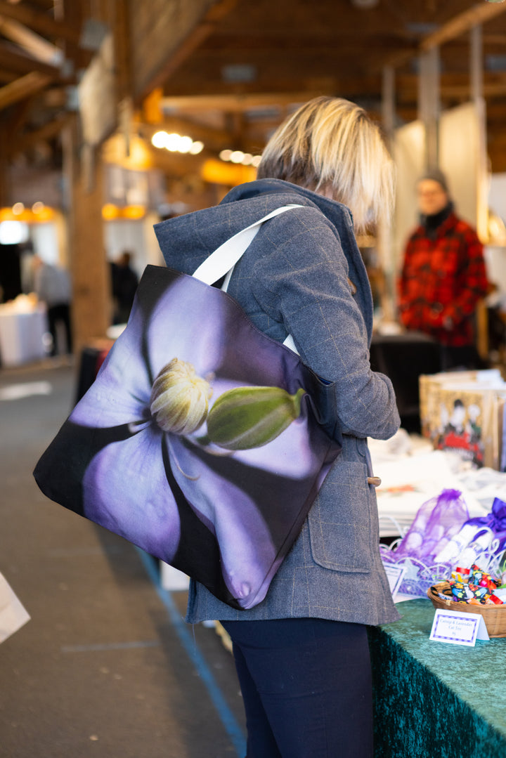 Woman at an indoor market carrying a tote bag with a soft purple clematis flower design on a dark background