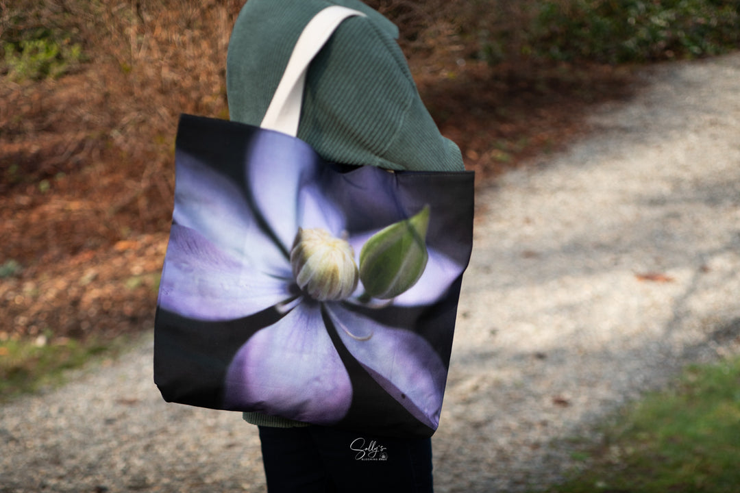Person walking along a wooded path carrying a tote bag with a soft purple clematis flower design on a dark background.