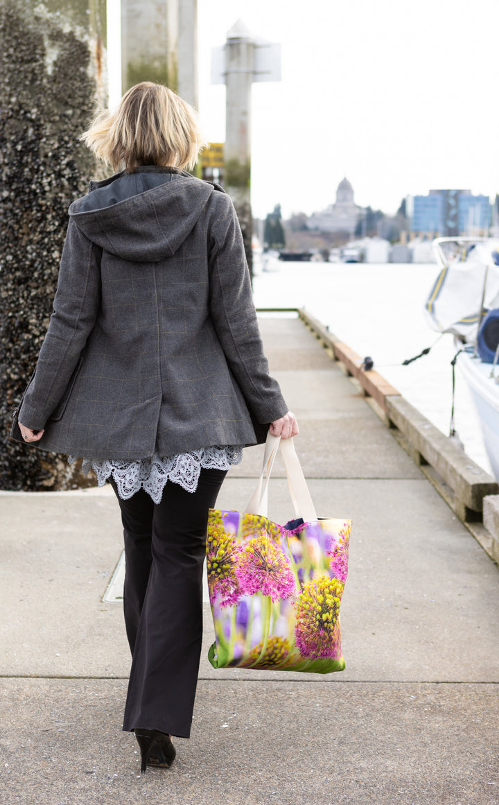 Woman walking along a waterfront walkway carrying a floral tote bag with a purple and yellow allium design.