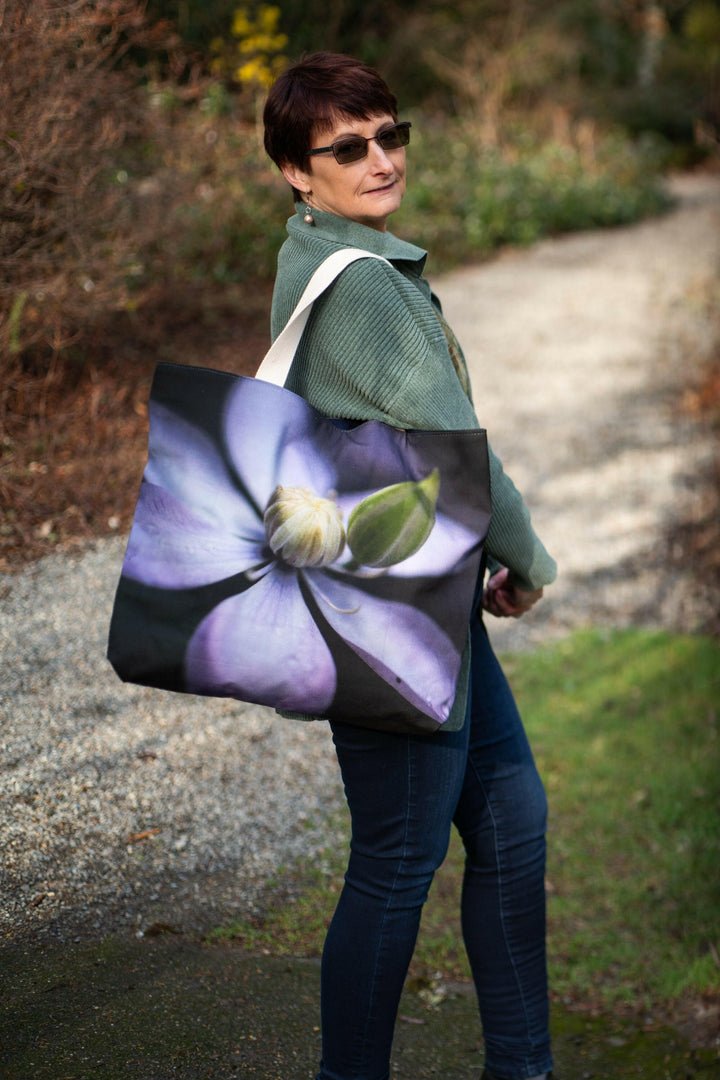 Woman walking along a garden path carrying a tote bag with a soft purple clematis flower design on a dark background